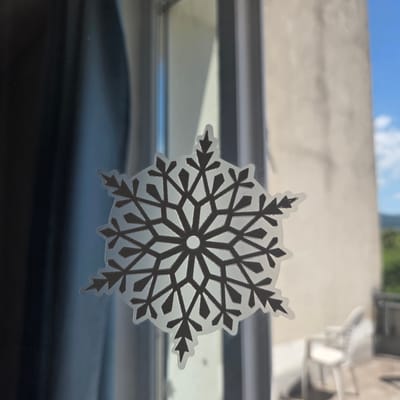 A close-up shot shows a snowflake-shaped sticker on a glass surface, likely a window. The sticker is detailed with intricate geometric patterns in a dark color against a translucent background, and it is outlined with a thin white border. Through the glass, a blurred background is visible, featuring a pale beige wall, a bright blue sky with scattered white clouds, and hints of greenery in the distance. To the right, a white plastic chair is slightly out of focus, suggesting an outdoor seating area. The overall impression is one of a decorative element on a window with an outdoor view. The image does not contain any discernible information that would place it specifically in the city of Piešťany, Slovakia.