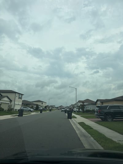 The image depicts a suburban street in Poinciana, Florida, United States, on an overcast day. The street is lined with modern residential homes, many of which appear to be two-story structures with garages. Cars are parked in driveways and along the street. Several black trash cans are visible on the curbside in front of the houses. The sky is heavily clouded, casting a diffused light over the scene. The environment is residential, typical of a planned community. There are no people visible in the image.