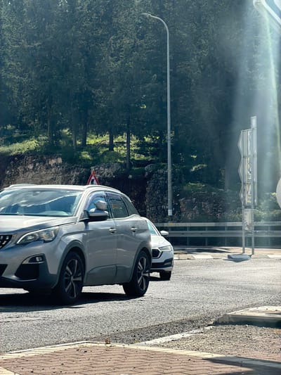 The scene depicts an outdoor roadway within the Merom HaGalil Regional Council, Israel, under bright daylight conditions with prominent sun flares originating from the right. A silver Peugeot SUV, likely a 3008 or 5008 model, is positioned in the immediate foreground on a paved asphalt surface, oriented slightly towards the right. Partially visible behind it is the front section of a white sedan. The roadway exhibits white painted lines and a curb transitioning to a brown brick-patterned pedestrian surface in the lower left. To the left of the road, a steep embankment rises, densely covered with green coniferous trees and interspersed with rocky outcrops. A triangular warning sign with a red border and a black pedestrian silhouette is partially visible on this embankment. A tall, grey streetlamp with an LED luminaire is situated on the right side of the road. Further right and behind the vehicles, a vertical support post holds multiple reflective grey road signs, and a silver metal guardrail runs parallel to the road. No individuals are present in the visible area.