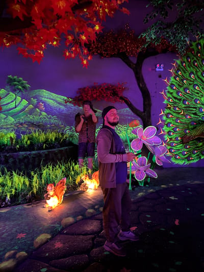 A man and a woman stand in an environment with fluorescent lighting and painted walls. The man wears a hat, jacket, and pants, and holds purple flowers with glowing accents. The woman stands slightly behind him. Behind them, a wall is painted with a nature scene including trees, flowers, and a peacock. Along the ground, there are additional painted elements and lit figurines. The location is a UV-lit mini golf venue in Binh Lu, Vietnam.