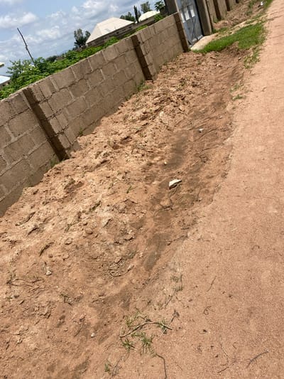 The image depicts an outdoor scene in Tavo, Nigeria, on a sunny day. The primary subjects are a wall constructed from concrete blocks, a sandy, dirt path, and sparse vegetation. The wall runs diagonally across the bottom left of the frame, suggesting a boundary or structure. The path, which occupies the majority of the image, is a mix of reddish-brown dirt and sand, with some patches of damp earth and dried debris.

Small tufts of green grass and dry, twig-like vegetation are scattered throughout the path, indicating a semi-arid or recently disturbed environment. In the background, visible beyond the wall, are hints of buildings, trees, and a cloudy blue sky. The lighting suggests it is daytime, likely midday or early afternoon, given the strong shadows and bright illumination. There is no discernible activity or event taking place; the scene appears static. The overall impression is of a rural or semi-developed area undergoing some form of construction or maintenance, given the presence of the block wall. No text is visible in the image.