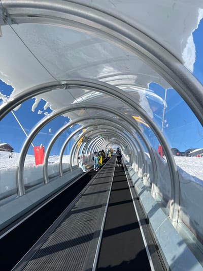 This vibrant image depicts the interior of a transparent, enclosed conveyor belt ski lift, often referred to as a "magic carpet," at a ski resort in Val-d'Isère, France. The perspective looks down the long, tubular structure, which is framed by repeating silver-colored metal arches and clear panels, creating a strong sense of depth.
Several individuals in winter ski attire, including one person in a prominent yellow jacket, are visible on the black conveyor belt, being transported uphill. Some are on skis, while others are standing. To the right of the moving belt, a stationary, textured black surface serves as a pathway or queuing area.
The transparent walls offer panoramic views of the bright blue sky and the surrounding snow-covered mountain landscape. Patches of snow cling to the exterior and interior edges of the transparent panels, particularly on the left side, indicating the alpine environment. The scene is brightly lit by natural sunlight, suggesting a clear, sunny day, with distinct shadows cast on the conveyor belt and the snow outside the tunnel. A red sign with illegible white text is partially visible outside the tunnel structure, likely indicating a safety message or information. The image captures modern ski infrastructure designed for convenient and scenic ascent in a snowy mountain setting.