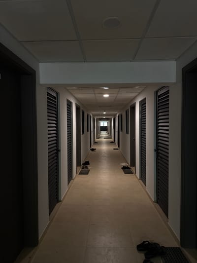 The image shows a long, empty hotel corridor in the Maldives. On either side of the corridor are closed doors with louvered panels, suggesting they lead to hotel rooms. Various pairs of sandals and flip-flops are placed on small mats outside some of the doors. The floor is tiled with large, light-colored square tiles. The ceiling is made of suspended acoustic tiles with recessed lighting fixtures. The corridor extends into the distance, with more doors visible on both sides. At the far end of the corridor, a brightly lit exit sign is visible. The scene is well-lit, with a natural light source at the end of the corridor and artificial lighting from the ceiling fixtures. The overall impression is one of cleanliness and order, typical of a hotel environment.