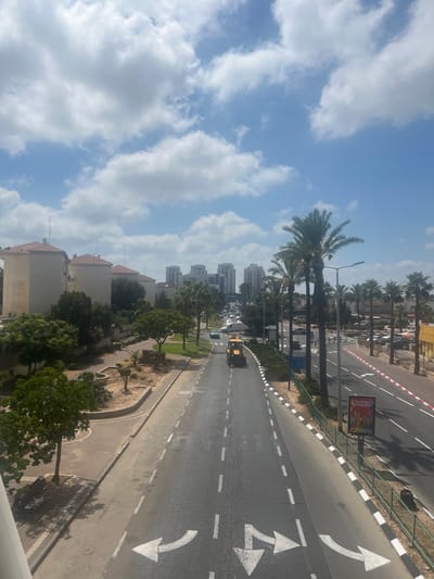 The image depicts an aerial view of a street scene in Or Akiva, Israel. The perspective is from a high vantage point, looking down at a road that curves to the right, lined with palm trees and low-rise buildings on one side and a sidewalk and road on the other. The sky is a vibrant blue, dotted with fluffy white clouds, suggesting a bright, clear day.

On the road, a yellow construction vehicle is visible, as well as other vehicles further down the street. The road is marked with white lines, indicating lanes and directional arrows. A billboard with colorful graphics and text is placed along the roadside. The buildings in the foreground appear to be residential, with tiled roofs and light-colored facades. In the background, taller, modern buildings rise against the sky. Lush green trees and landscaping are present along the sides of the road and in front of the buildings, adding a touch of nature to the urban environment.

The scene suggests a typical day in a city or town. The bright sunlight casting shadows indicates it is likely daytime, possibly late morning or early afternoon. The overall mood is one of everyday activity and a pleasant, sunny atmosphere. The visible text on the billboard is in Hebrew, which, along with the location context of Or Akiva, Israel, confirms the geographical setting.