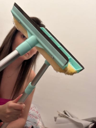 A woman is reflected in a mirror as she holds a window cleaning tool. The tool has a light green handle and frame, a yellow sponge, and a black rubber squeegee. She is holding the handle with her right hand. Her long, dark hair is draped over her shoulder. A white wall is visible in the background, along with a partial view of clothing.