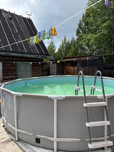 In Zheleznodorozhnyi, Russia, a backyard scene is depicted with an above-ground swimming pool as the central subject. The pool is filled with green water and has a tiled interior pattern. A metal ladder with white steps provides access to the pool. The pool is situated on a wooden deck next to a rustic wooden building with a dark, weathered roof. A clothesline stretches across the scene, adorned with several colorful clothespins. Trees and other wooden structures are visible in the background. The sky is overcast. There are no people present in the image.
