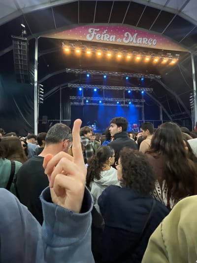 The image captures a scene from the "Feira de Março" event in Aveiro, Portugal. A crowd of people is visible, mostly looking towards a stage illuminated with bright orange and blue lights. A banner with the event's name is visible on the left. The photo is taken from within the crowd. A person in the foreground is pointing a finger towards the stage. The environment appears to be an indoor event tent or similar covered venue.