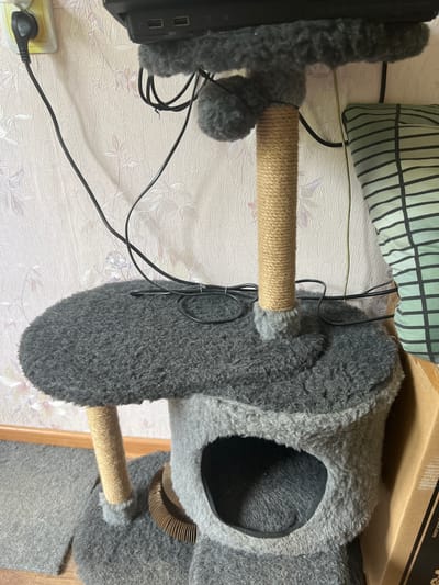 The image shows a cat tree in an indoor setting. The cat tree is gray and made of plush material and rope. It has multiple levels, including a platform and a cubbyhole. Wires are visible around the cat tree and plugged into an electrical outlet on a wall with floral wallpaper. A pillow with a striped pattern is on the right side of the image, and a cardboard box is partially visible. The scene depicts a domestic environment.