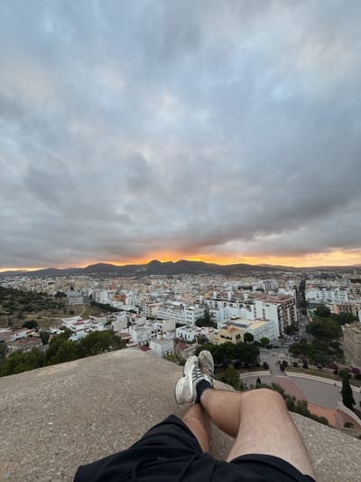 A low-angle, first-person perspective photograph captures the lower legs of a person resting on a rugged, light grey concrete or stone surface, overlooking the city of Eivissa, Spain. The person wears black shorts, white socks, and worn white sneakers with black stripes. Below, the city sprawls with dense clusters of white and light-colored buildings, ranging from residential houses to multi-story structures, intersected by roads and green areas. Distant hills form the horizon line. The sky is overcast with grey clouds, but a vivid band of orange and yellow light from the setting sun is visible along the horizon, illuminating the clouds from beneath and casting an evening glow over the urban landscape. The scene depicts a tranquil observation of the sunset over the city.