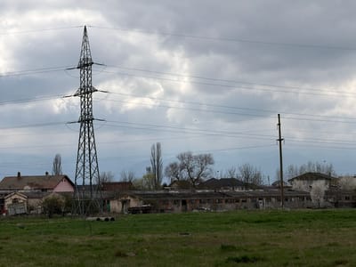 The image depicts a rural scene in Sibiu, Romania, under an overcast sky. In the foreground is a field of green grass. A metal power line tower stands prominently in the left-center of the frame, with overhead power lines stretching across the sky. Behind the tower, there are old, dilapidated buildings, including residential houses and what appear to be abandoned agricultural structures. Trees with bare branches are interspersed among the buildings. The background includes a hazy mountain range. The overall impression is one of decay and rural poverty.