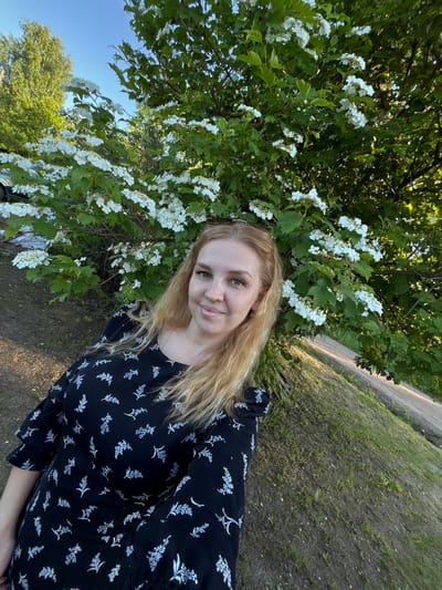 The image captures a fair-skinned woman, appearing to be in her late 20s to early 40s, with long, straight light blonde hair and light-colored eyes. She has freckles on her nose and cheeks and is smiling subtly while looking directly at the camera. She wears a black dress with a repeating pattern of white floral or botanical motifs, and the sleeves have ruffled detailing. A thin, gold-colored chain is visible around her neck. The woman is positioned at an angle in the lower-left and central portion of the frame, appearing to take a selfie.

She is standing outdoors beside a prominent bush laden with clusters of small white flowers and green leaves, consistent with a species like Viburnum. This flowering bush dominates the central and right side of the background. The ground immediately around her and extending to the left is unpaved, composed of dark earth with sparse green grass and weeds. In the background, beyond the flowering bush, there are larger green trees and a clear, bright blue sky. A dirt road or unpaved track is visible horizontally across the mid-ground on the right side of the image. A light-colored vehicle is partially obscured by foliage in the distant left background. The scene is illuminated by natural sunlight, indicating either late morning or late afternoon. The location is Urmanets, Russia.