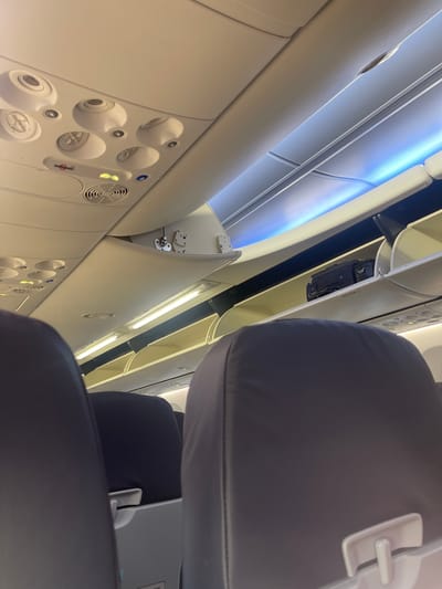 The image displays the interior of a commercial aircraft cabin, observed from a passenger's perspective, angled forward and upward. Two dark-colored passenger seatbacks are partially visible in the immediate foreground. Above, the overhead panel features multiple circular air vents, individual reading lights indicated by lightbulb icons, an illuminated red "No Smoking" sign, an illuminated yellow "Fasten Seatbelt" sign, and a blue "Flight Attendant Call" button. A circular grille is also present on this panel. Overhead storage bins line both sides of the cabin. On the right side, several bins are open, and a small black, rigid case or bag is visible inside one of them. Cabin lighting consists of linear white lights beneath the overhead bins and a strip of soft blue ambient lighting further up on the ceiling, above the bins on the right. No individuals are visible within the frame. Given the provided information, the location is associated with Crawley, United Kingdom.