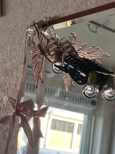 The image shows a close-up of a decorative mirror with various ornaments. A pink ribbon is tied in a bow to the left side of the mirror frame, with the ribbon extending downwards. Attached to the top of the mirror are metallic ornaments resembling insects or mythical creatures, with delicate wings and intricate details. Small, clear bulb lights are strung along these ornaments. The mirror reflects a room with textured walls and a window with a view of buildings. The location is not specified in the image or its context, therefore it cannot be determined to be Genemuiden, Netherlands.