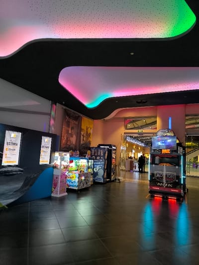 The image captures the vibrant interior of an entertainment venue, likely an arcade or a section within a larger complex such as a cinema or shopping mall, situated in Agios Dimitrios, Greece. The space features a dark, reflective tiled floor that mirrors the colorful lights throughout the room.

A dominant visual element is the modern ceiling, constructed from undulating white panels with small perforations, dramatically lit by hidden LED strip lights. These lights cast dynamic washes of pink, purple, and green hues onto the ceiling, creating an energetic ambiance. The ceiling's curves are accentuated by dark, recessed borders.

On the left side of the frame, two tall, dark digital screens display Greek text "Εισιτήρια" (Tickets) and "ΣΤΟ ΚΙΟΣΚ" (To the Kiosk), indicating a ticket purchasing station. Below these screens, a dark, sloped surface with a bright blue edge extends into the foreground. Alongside, several arcade game machines are visible, including brightly colored claw machines filled with toys and another blue machine with visible text referencing "FACEBOOK".

Towards the right, a prominent motion simulator arcade game, labeled "MOTION PARK" and "RACE THE...", stands out. This game features a screen and a driver's seat, casting distinct red and blue light reflections onto the tiled floor. In the background, a large poster with the Greek text "Υπάρχω" (I Exist) is displayed.

Through an archway in the far background, a few indistinct people can be seen near an "EXIT" sign, hinting at further areas or an exit from the complex. Decorative string lights are also visible in this background section, adding to the festive atmosphere. The artificial lighting suggests an indoor setting, likely conducive to entertainment regardless of the time of day, but often associated with evenings. The overall scene conveys a lively leisure environment designed for amusement.