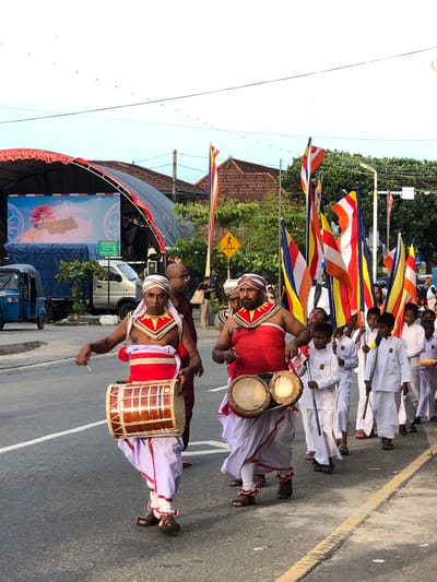In Habaraduwa, Sri Lanka, a procession moves along a paved road under a bright sky with scattered birds. In the foreground, two men, dressed in traditional white and red ceremonial attire with elaborate headbands and chest adornments, are playing drums. The man on the left holds a large, cylindrical drum horizontally, while the man on the right plays a set of two smaller, hourglass-shaped drums suspended from his waist. Both men are actively engaged in drumming, with sticks in hand.
Behind them, a group of boys, also in white attire, follow carrying long sticks, possibly part of a ceremonial guard or participants. In the background, several Buddhist flags, featuring stripes of orange, yellow, red, and blue, are visible, adding to the festive or religious atmosphere. Beyond the procession, a stage structure with a large screen displaying an image of a lotus flower is set up, with a blue truck and a small three-wheeled vehicle parked nearby. Buildings with tiled roofs and trees line the side of the road, indicating an urban or semi-urban setting. The overall scene suggests a cultural or religious event taking place in the town.