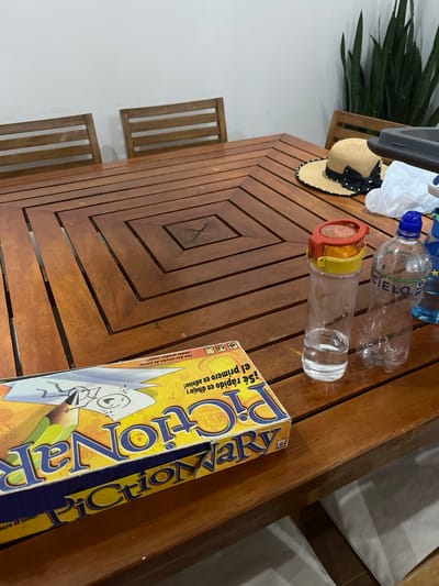 The image shows a wooden table with a slatted surface. On the table, from left to right, are: a potted plant with long, green leaves; a straw hat with a black ribbon; a plastic bag; two partially empty plastic water bottles, one labeled "CIELO"; a water bottle with an orange cap and a yellow band; and a Pictionary game box in Spanish. The table is set against a white wall and part of the leg and seat of a chair can be seen on the left and partially on the right of the table. The image's environment suggests an indoor or enclosed space. The location appears to be Pacarán, Peru, based on the language on the game box.