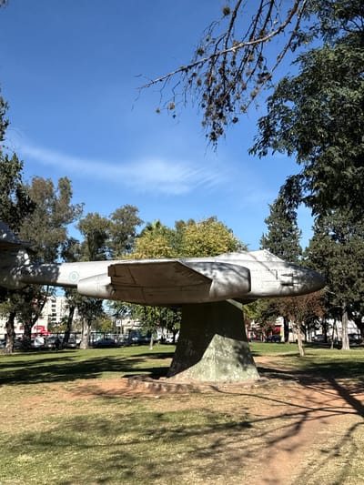 The image shows an IA-58 Pucará aircraft monument located in the city of Salta, Argentina. The aircraft is mounted on a concrete plinth and is positioned on a grassy area surrounded by trees and buildings. The sky is visible in the background, and the monument is casting a shadow on the ground.