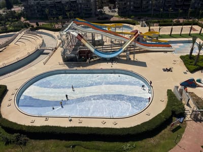The image, taken in Erdemli, Turkey, shows an aerial view of a water park under maintenance. The main pool is oval-shaped and empty, with workers cleaning its tiled surface. The pool is patterned with alternating light and dark blue tiles. A row of small concrete steps and platforms encircle the pool.
Several water slides of various colors (yellow, blue, orange, white, and red) are present. A building labeled "ül-fet SNACK BAR" is visible beneath the largest water slide structure. The area surrounding the pool and slides is mostly paved.
There are sections of landscaping, including grass, palm trees, and manicured hedges. A multi-story residential building is in the background.
Several individuals are cleaning the pool using tools such as brushes and buckets.