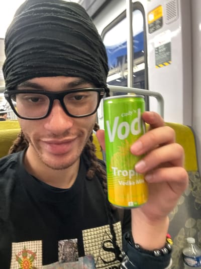 A person is holding a can of “Cutwater Vodka Mix Tropical” inside what appears to be a train car. The person has dark hair partially covered by a black head wrap, and they are wearing black-framed glasses and a black shirt with patches. The can is green and yellow, with the words “Cutwater Vodka Mix Tropical” prominently displayed. A plastic water bottle is visible in the background. The location is a train car, potentially in the city of Arcueil, France. The train car contains yellow seats, and signs with information for passengers.