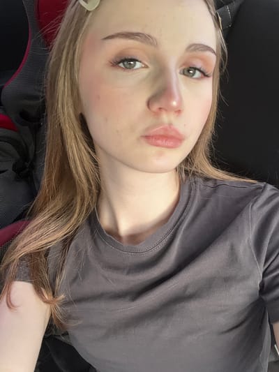 A young woman with blonde hair is captured in a close-up, head-and-shoulders shot. She is looking directly at the camera with a neutral expression. Her eyes are a hazel green, and she is wearing subtle eye makeup. Her lips are slightly parted, and her skin appears smooth with a touch of blush on her cheeks. She has a light-colored hair clip visible in her hair. She is wearing a plain, dark gray t-shirt. The background is a mix of a dark, possibly upholstered surface and a black background, suggesting she might be seated in a chair or in a vehicle. The overall lighting is soft, highlighting her facial features. The image does not contain any information that would allow for its location to be determined within the city of Новая Отрадовка, Russia.