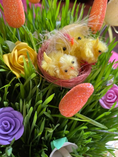 A close-up shot displays a nest filled with fuzzy, yellow chicks amidst lush, green grass. Several orange, glittery carrots and colorful roses are scattered around the nest and grass. The roses are pastel shades of purple, yellow, and pink. The scene evokes a spring or Easter theme. Location information (Aratashen, Armenia) is not visually discernible in the image.