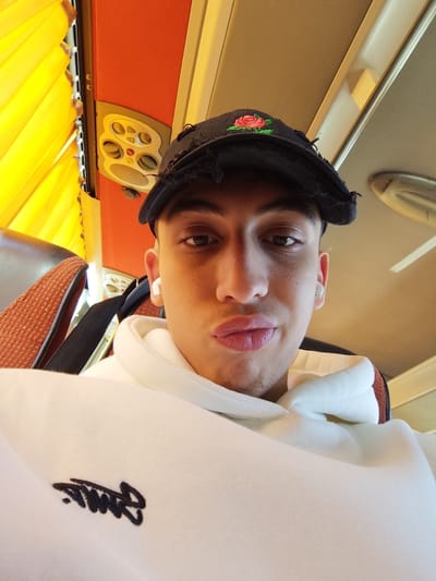A young male is depicted in a close-up, selfie-style photograph taken inside what appears to be a passenger bus or coach in Jibb as-Safa, Syria. The individual has a fair complexion, short dark hair, and is wearing a distressed black baseball cap with a red and green rose embroidery on the front. He is attired in a white hoodie featuring a black script logo on the left chest area. White wireless earbuds are visible in both ears. He presents a pouting expression, looking directly into the camera. The background shows part of a brown and orange upholstered seat, an overhead panel with circular ventilation vents and controls, and yellow pleated window blinds, consistent with a vehicle interior.