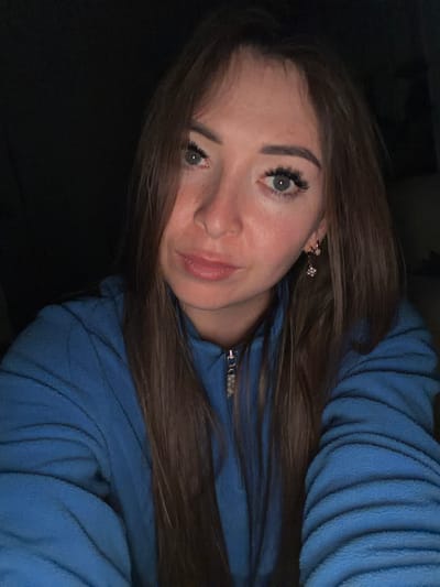 A young woman with long brown hair, fair skin, and light blue eyes is pictured from the chest up, looking directly at the camera. She is wearing a blue fleece jacket with a zipper. She has on delicate earrings, one visible, that appear to have a star-shaped charm. The lighting is dim, creating a dark background. The image does not contain any specific geographical markers or elements that would indicate a location within the city of Япрыково, Russia.