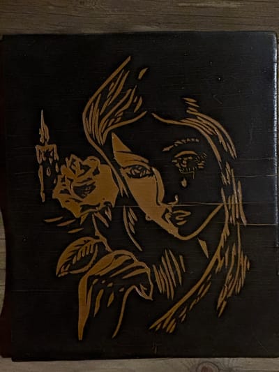 The image shows a dark, likely wooden, surface with a carved or engraved design. The design is in a lighter color, possibly exposing the natural wood beneath the dark surface. The design depicts a stylized woman's head and upper body with flowing hair, appearing to be under an umbrella or canopy. Below the woman, a rose blossom with leaves is included in the composition. The surface appears to be part of a piece of furniture, possibly a stool or box. The background is out of focus, showing a wooden floor.
 Маза, Russia, is not discernible from the image.