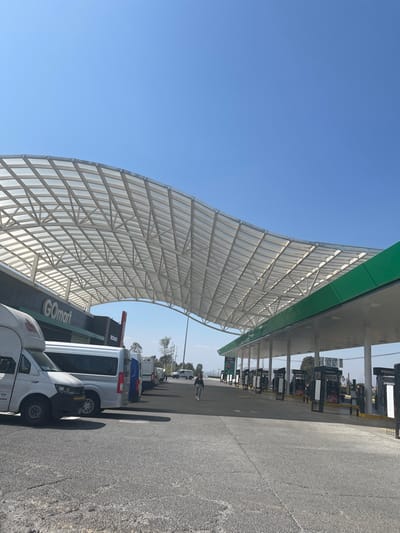 A broad view of a gas station on a clear day. The station features a vast, white grid-like canopy providing shade over multiple gas pumps. A GoMart convenience store is visible, with several white vans parked nearby. A cyclist is positioned near the center of the shot. The sky is a clear, bright blue, contrasting with the gray pavement and vehicles.