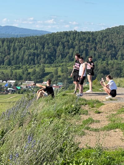This image captures a scenic overlook in the city of Sim, Russia, on a bright, sunny day. In the foreground, lush green grass and wild purple flowers grow on a gentle slope. A man sits on the grass, pointing towards the valley below, which features a village with houses and a road with trucks. Behind the village, a dense forest covers the rolling hills, with a clear blue sky overhead. On the dirt path to the right, four young adults are gathered. Two men and a woman stand together, while another woman crouches down, taking a photo with her phone. The scene evokes a sense of outdoor recreation and appreciation for the natural beauty of the region.