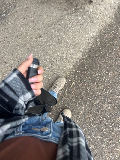 The photograph shows a person standing on a paved surface, likely a street or sidewalk, holding a black object, possibly a remote or electronic device, in their left hand. The person is wearing light blue jeans, beige sneakers, and a buttoned denim jacket over a brown top, and a gray and black plaid shirt over that. A black bag hangs across their body. The paved surface is gray with small pebbles and appears to be asphalt or concrete. The setting cannot be independently verified as Blizhniy Khutor, Moldova, due to a lack of geographical or contextual information within the image itself.