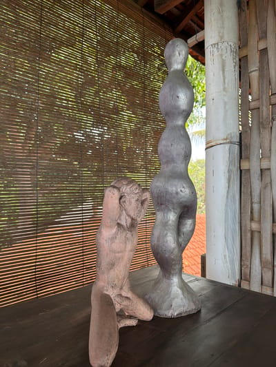 The image depicts a still life scene on a dark wooden table in Tejakula, Indonesia. Two sculptures are the primary subjects. To the left, a pinkish, textured sculpture of a reclining male figure is positioned, with his hands raised to his head. To the right, a taller, silver-toned abstract sculpture with a bulbous, undulating form stands upright. In the background, a bamboo blind covers a window, allowing dappled sunlight to filter through, creating a pattern of light and shadow on the scene. Beyond the blind, glimpses of lush greenery and a terracotta-tiled roof are visible. To the right of the sculptures, a white pillar and a vertical arrangement of bamboo poles form part of the architectural environment. The overall impression is one of an outdoor or semi-outdoor setting, possibly a patio or veranda, with natural elements integrated into the decor.