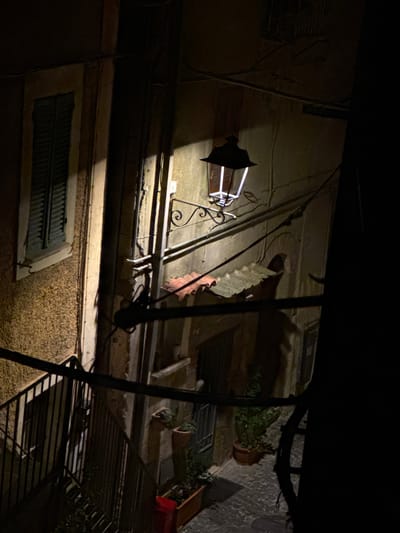 A nighttime shot, taken in Castelnuovo di Porto, Italy, of a building facade viewed from a low angle. The scene is predominantly dark, illuminated by an ornate, wall-mounted lamp with a white glow, casting light directly onto the building. Architectural features include windows with dark, louvered shutters, one directly below the light, and others higher up on the building. Electrical wires run along the walls. Potted plants sit on a windowsill. A metal structure is in the foreground. The street is cobbled.