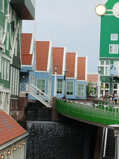 This image depicts a picturesque scene in Zaandam, Netherlands, showcasing charming, traditional Dutch-style buildings. The architecture features distinct green facades with white trim, and a row of pastel blue houses with orange-tiled roofs. A white staircase leads up to the blue houses, and a dark waterfall cascades below. A vibrant green bridge with white railings spans across the water. Several people are visible on the bridge, some walking, others standing and observing the surroundings.
The weather appears to be overcast, suggesting it might be a cloudy day. The time of day is not clearly indicated, but the lighting suggests daytime. A prominent feature in the background is a tall green building with a large circular clock face and a smaller round element above it, characteristic of some historical buildings in the region. A light blue swan-shaped decoration is visible on the side of this green building.
The scene conveys a sense of calm and leisurely atmosphere, with people enjoying the view of the unique architecture and the water feature. The overall impression is one of a tourist-friendly location with well-preserved historical architecture. No visible text is discernible in the image.