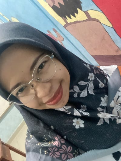 The image is a close-up, angled selfie of a young woman wearing glasses and a dark hijab with a floral pattern. The background appears to be a brightly painted mural on a wall, with blue and red sections and what seems to be stylized figures. A wooden railing is visible in the lower left corner, suggesting an indoor or covered outdoor setting, possibly a home or a communal area in Tanjung Gading, Indonesia. The woman's face is the primary focus, with her eyes looking towards the camera and her lips slightly parted in a gentle smile. The lighting is soft, highlighting her features and the details of her hijab.