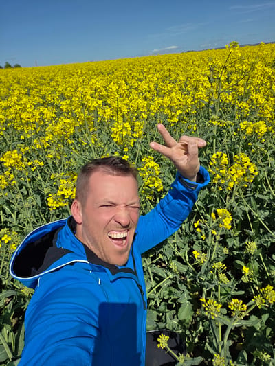 A man lies in a field of yellow flowers. He is fair-skinned, has short hair, and wears a blue hooded jacket. His mouth is open in a wide smile, and he holds up his hand, fingers forming a "peace" or "victory" sign. The field is filled with yellow flowers, contrasting with the clear blue sky.
The location is Ihringshausen, Germany.