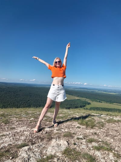 The image depicts a young woman standing on a rocky outcrop overlooking a vast, forested valley under a clear blue sky. She is wearing an orange crop top and a white denim skirt with frayed edges. Her left arm is outstretched horizontally to her side, and her right arm is raised vertically with her index finger extended. She is also wearing sunglasses and light-colored wedge sandals. The terrain in the foreground is characterized by scattered rocks and sparse grass. In the background, a dense forest covers the undulating landscape, with patches of green fields visible in the distance. The location is identified as the city of Shikhan, Russia, specifically on a high vantage point, likely a mountain or hill offering a panoramic view. The scene suggests a bright, sunny day suitable for outdoor activities and sightseeing.