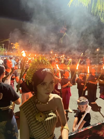 The image displays a nighttime procession occurring on a street in Munggu, Indonesia. The scene is primarily illuminated by numerous flaming torches carried by participants, generating a significant amount of smoke that creates a hazy, glowing atmosphere.
In the immediate foreground, a young adult female is positioned slightly right of center, facing partially right. She is adorned in traditional ceremonial attire, including a gold ornate crown, a one-shoulder garment made of striped or patterned fabric, a prominent gold broach on her shoulder, a pearl necklace, and a gold bangle on her left wrist. Her expression appears composed.
Behind her, a dense crowd of individuals, comprising both adults and children, fills the paved street. Most participants are holding large bamboo torches with active flames. Several children are dressed in dark shirts and traditional sarongs, with one small child in the lower right foreground wearing a headwrap and holding a white drumstick next to a partially visible dark object. To the mid-left, an adult female is visible with an open mouth, suggesting vocalization.
In the background, tall palm trees are present. On the far left, a large, elaborate effigy or sculpture stands, partially obscured by the crowd and smoke. On the far right, a stone architectural element, consistent with a temple gate, is discernible. The overall environment depicts a lively cultural or religious event.