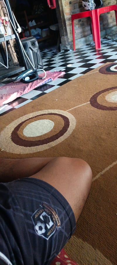 The image shows a low-angle perspective of a person's legs and lower torso, partially visible on a brown carpet with concentric circle patterns. The person is wearing dark athletic shorts with a crest featuring a soccer ball and some text. The background includes a black and white checkered floor, a pink ironing board with a red iron on it, a large black speaker, and a red plastic chair. The setting appears to be indoors, possibly a living room or a covered outdoor area. The brick wall texture in the background suggests a rustic or traditional building style, common in areas like Kemuning, Indonesia. The lighting is natural and diffused, indicating daytime.