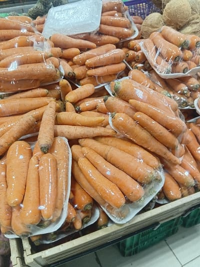 The image shows a large quantity of carrots arranged in plastic-wrapped packages, stacked on top of each other and displayed on a wooden shelf in a grocery store. The carrots are orange in color, some are longer and thinner than others. The shelf is made of wood with a green plastic crate underneath. The image was taken in a grocery store at the location of coordinates: latitude: -1.417, longitude: 36.953, which is in Pridelands, Kenya.  No people are present.  The store is well lit.