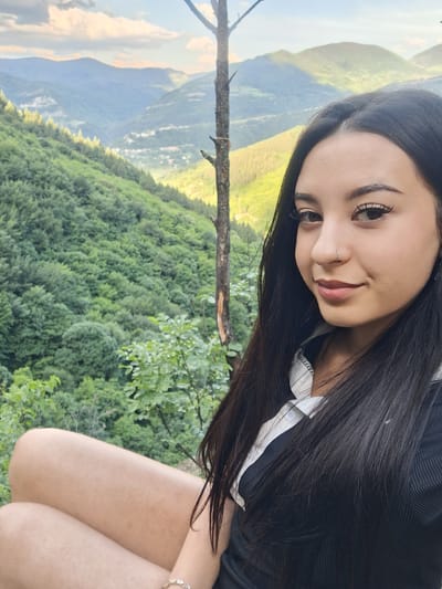A young woman with long, dark hair poses in front of a lush green mountain landscape. She has fair skin and a nose ring. She is wearing a black shirt with a white collar underneath. Part of her leg is visible in the lower right corner of the frame. The landscape behind her features mountains, trees, and a wooden branch extending into the frame. The image appears to be taken in the Zasele, Bulgaria region.