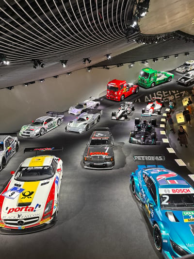 This image captures an indoor exhibition of various Mercedes-Benz racing cars and trucks. The scene appears to be the Mercedes-Benz Museum in Stuttgart, Germany, judging by the vehicles and overall presentation. The exhibition is set against a modern, minimalist backdrop with a slatted ceiling and track lighting.
In the foreground, a white and yellow racing car with "porta!" and "BLACK FALCON" branding is prominently displayed. To its right, a bright blue racing car with "Mercedes-Benz" and "BOSCH" logos is visible. Further up and to the left, a silver racing car with "WARSTEINER" branding is parked.
Behind these, a collection of other racing cars and trucks are arranged. These include a red truck with "ESSO" branding, a green truck with "BP" livery, several silver and white Formula 1-style cars, and a black racing car with "SONAX" branding. In the background, a few visitors can be seen observing the exhibits. The floor is a dark, matte surface, contributing to the sophisticated presentation of the vehicles.