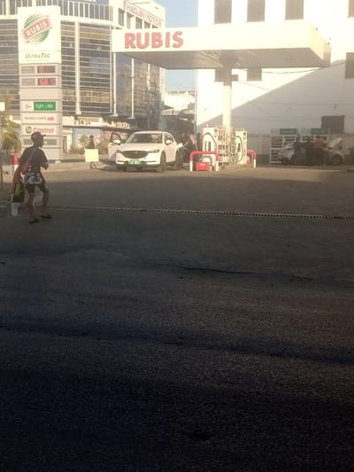 The image depicts a daytime scene at a Rubis gas station in Ganjoni/Shimanzi ward, Kenya. In the foreground, a dark-skinned individual wearing a dark t-shirt, patterned shorts, and sandals walks rightward, carrying a red bag and a white bottle. To the left, a tall digital price display sign branded "RUBIS UltraTec" shows fuel prices, including "168.10," and logos for "light.zille" and "Castrol." Another person stands near this sign, holding a white rectangular object.
At the fuel pumps, a white Mazda SUV with a green license plate (partially visible as "K... 06L") is being refueled by an attendant, with its driver's side door open. Further right under the white Rubis canopy, two more light-colored vehicles are present; one has its hood open, and several individuals are gathered around them. The background features multi-story commercial buildings, one displaying "HAOJIN MOTOR" signage. The scene is illuminated by strong sunlight, causing the immediate foreground to be in deep shadow, while the mid-ground and background are brightly lit. The ground is paved with dark asphalt.