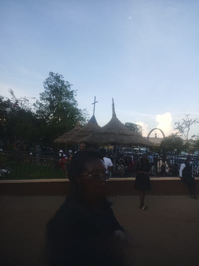 This image depicts an outdoor gathering in the Thika Township ward, Kenya, during either dusk or dawn. The foreground features an individual with dark skin, wearing glasses and dark clothing, standing with arms crossed, partially obscured by shadow on the left. In the midground, two circular thatched-roof structures are visible; the left structure is topped with a cross. A black metal fence separates the foreground from a crowd of approximately 20-30 individuals gathered around and behind the thatched structures. A woman in a black dress stands prominently in the mid-right, while other people, including a man in a white shirt and another in a dark vest, are visible standing and seated. Vehicles are parked further in the background. The sky is a light blue, with a faint moon visible in the upper right quadrant. Trees frame the scene on the left and right, and a distinct white cloud formation is present in the far-right background, above an arched structure. The ground appears to be a mix of paved and unpaved areas.