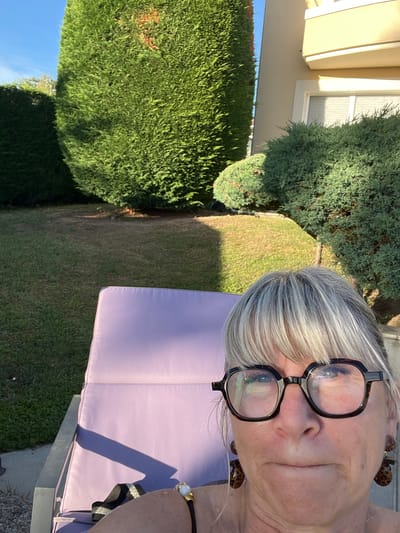 A selfie is taken from a low angle, capturing the face of a woman with blonde and grey hair and bangs, wearing large, dark-rimmed glasses and leopard-print earrings. She is looking directly at the camera. Behind her, a portion of a lavender lounge chair is visible, and further back, a manicured garden with tall, dense green hedges and a grassy area. Sunlight casts shadows across the scene, suggesting it is a bright, sunny day. The background also includes a portion of a building with a yellow facade and a balcony. The setting appears to be a private garden or patio area. The image was taken in Saint-Galmier, France.