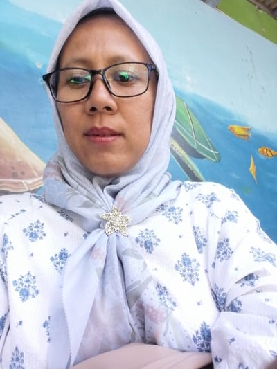A selfie taken by a woman wearing a hijab and glasses in front of a mural. The woman is wearing a white shirt with blue floral patterns and a light blue hijab with a floral pattern. She is also wearing a silver brooch shaped like a star on her hijab. The mural behind her depicts an underwater scene with a blue background, a green boat, and yellow fish. The location is Tegalwangi, Indonesia.