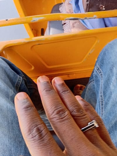 The scene captures a first-person perspective from within a bright orange/yellow vehicle, consistent with an auto-rickshaw, situated in Dorayi, Nigeria. In the foreground, a person's hand with dark skin pigmentation is prominent, displaying four visible fingers. A silver-colored ring with a black rectangular inlay is present on the ring finger. The person's lower body is clad in blue denim jeans. The vehicle's interior features orange metal structural components, including two horizontal bars and a lower panel. In the upper background, remnants of light blue fabric and a partial view of a bare foot are discernible, indicating another presence or interior element. The orange surfaces exhibit minor scuffing and paint wear.