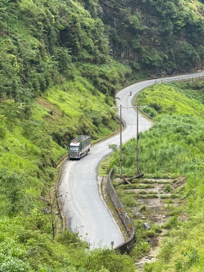 A bird's-eye view captures a winding road carved into the mountainside near the city of Yên Minh Commune, Vietnam. Lush green vegetation covers the slopes, with a grey concrete road cutting through. A truck navigates a curve in the road, while a lone figure on a motorcycle can be seen further up the path. Utility poles line the roadside, and stone retaining walls are visible on the steeper sections of the terrain.