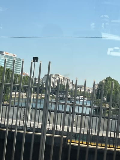 The image captures a sunny day with a clear blue sky over the city of Neuilly-sur-Seine, France. In the background, modern buildings with reflective windows are visible, some featuring logos. A body of water, likely the Seine River, separates the foreground from the buildings. The foreground is dominated by a metal fence, composed of vertical bars and horizontal railings, obscuring a portion of the view. Beyond the fence and the water, lush green trees line the riverbank. The scene appears to be taken from a slightly elevated position, possibly a bridge or an upper floor, looking across the river. There are no people or active events depicted in the image; it is a static, urban landscape.