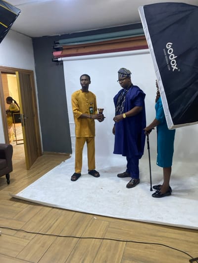 A photo shoot is in progress. Two men and a woman stand against a white backdrop, illuminated by a Godox softbox. The man in the foreground wears a mustard-yellow suit, holding a device. Behind him, a man in a blue robe is wearing a traditional Yoruba hat. The woman is wearing a turquoise skirt suit and holding a black cane. A door stands ajar, with another figure visible within. The room features a wooden floor and studio lighting equipment.